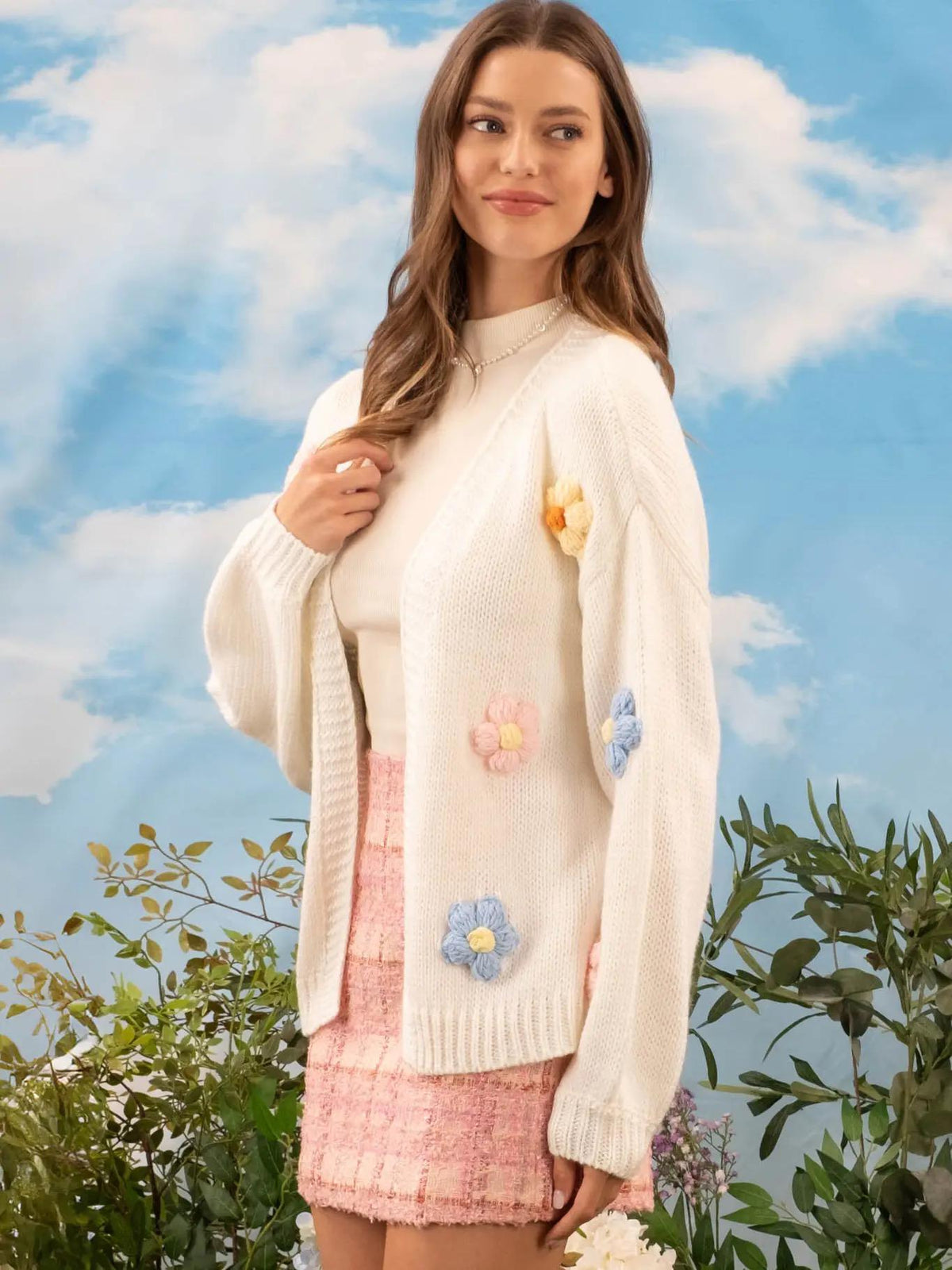 Woman wearing a white cardigan with floral embellishments against a blue sky and greenery background