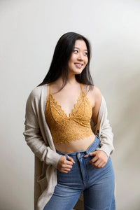 Woman wearing a yellow lace top and blue jeans with a beige cardigan against a plain background