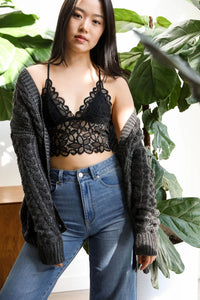 Woman wearing a black lace top and blue jeans with a gray cardigan, standing in front of green plants.