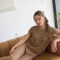 Woman sitting on a couch wearing a brown sweater and beige pants in a bright room.