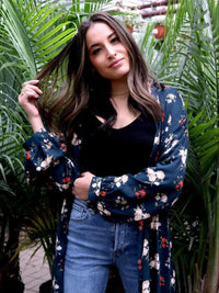 Woman wearing a floral kimono over a black top and blue jeans, standing in a tropical setting with palm leaves.