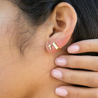 Close-up of an ear wearing a gold square hoop and gold cross earring with a hand touching the earlobe.