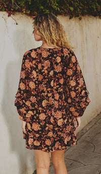 Woman wearing a floral dress against a white wall with greenery.