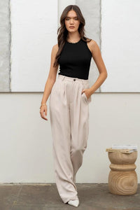 Woman wearing a black sleeveless top and beige high-waisted pants standing against a white wall.