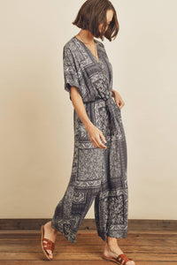 Woman wearing a patterned jumpsuit standing against a plain wall.