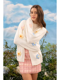 Woman wearing a white floral-patterned cardigan against a blue sky and green foliage background