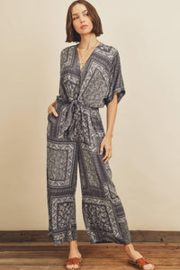 Woman wearing a patterned  grey jumpsuit standing against a plain background