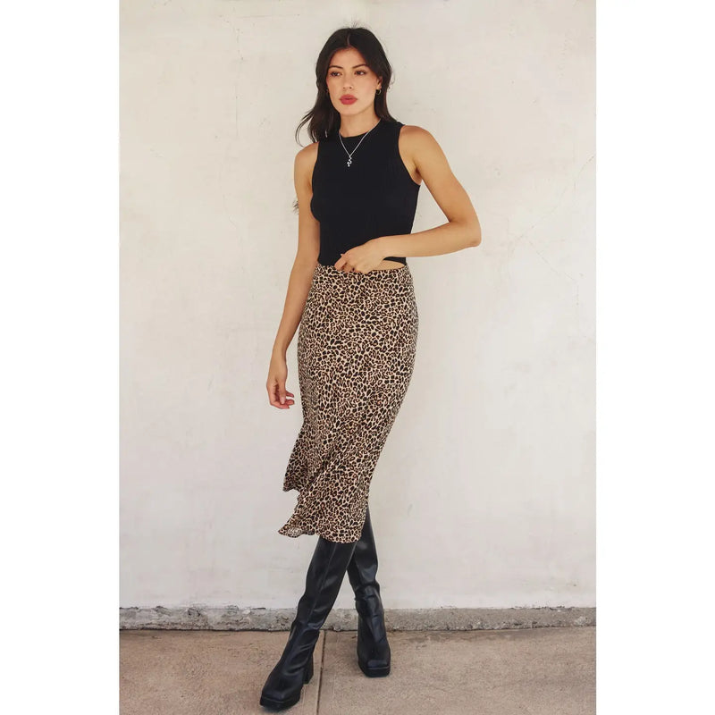 Woman wearing a black sleeveless top and leopard print skirt with black boots against a white wall.