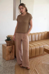 Woman wearing a brown top and beige pants standing in a living room with a beige sofa and wooden coffee table.